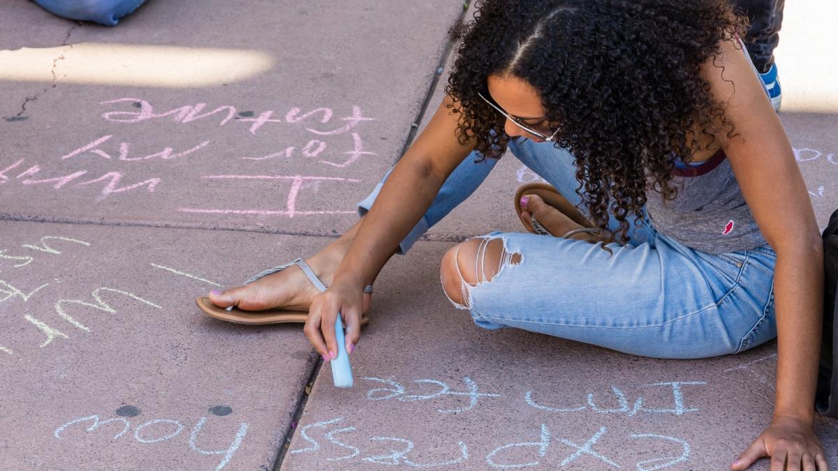 Female student siting on concrete, writing with chalk.